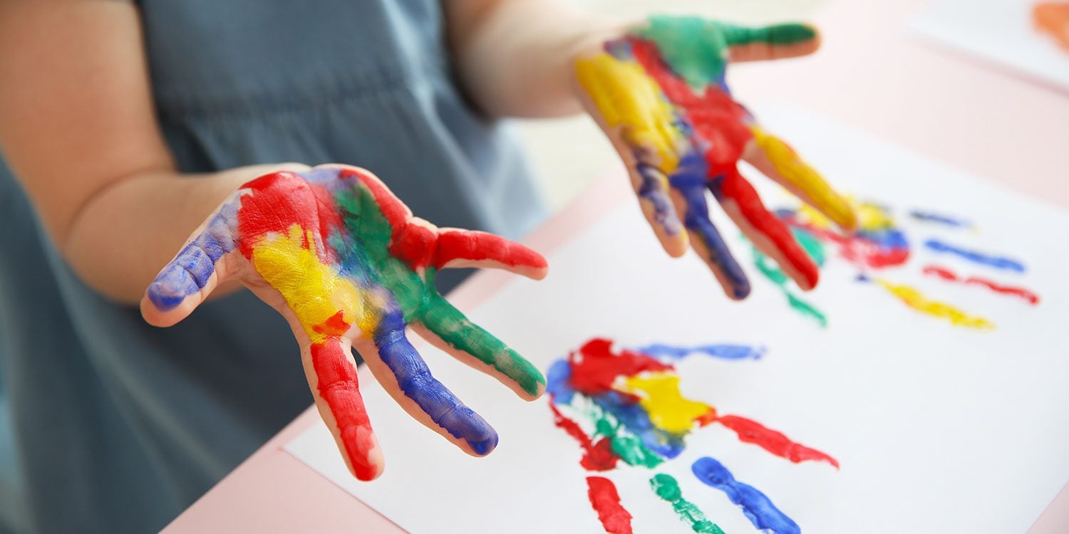 Child's hands covered in colorful paint, making handprints on paper.