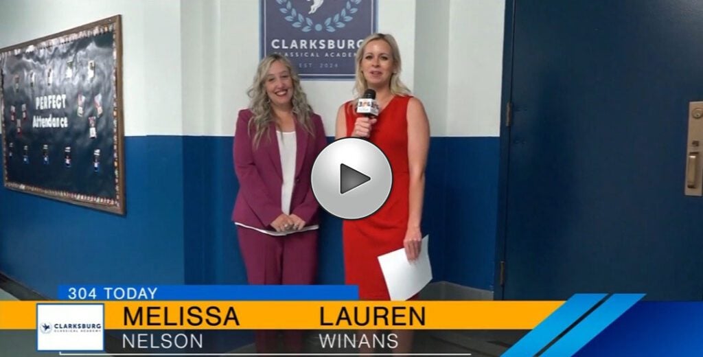 Two adults in a hallway, one with a microphone, near a "Perfect Attendance" board.