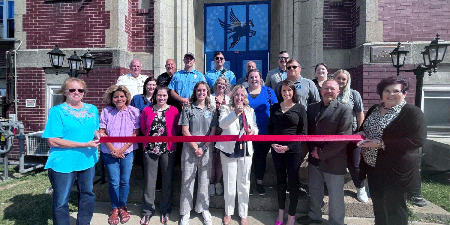Adults hold a red ribbon in front of a building.