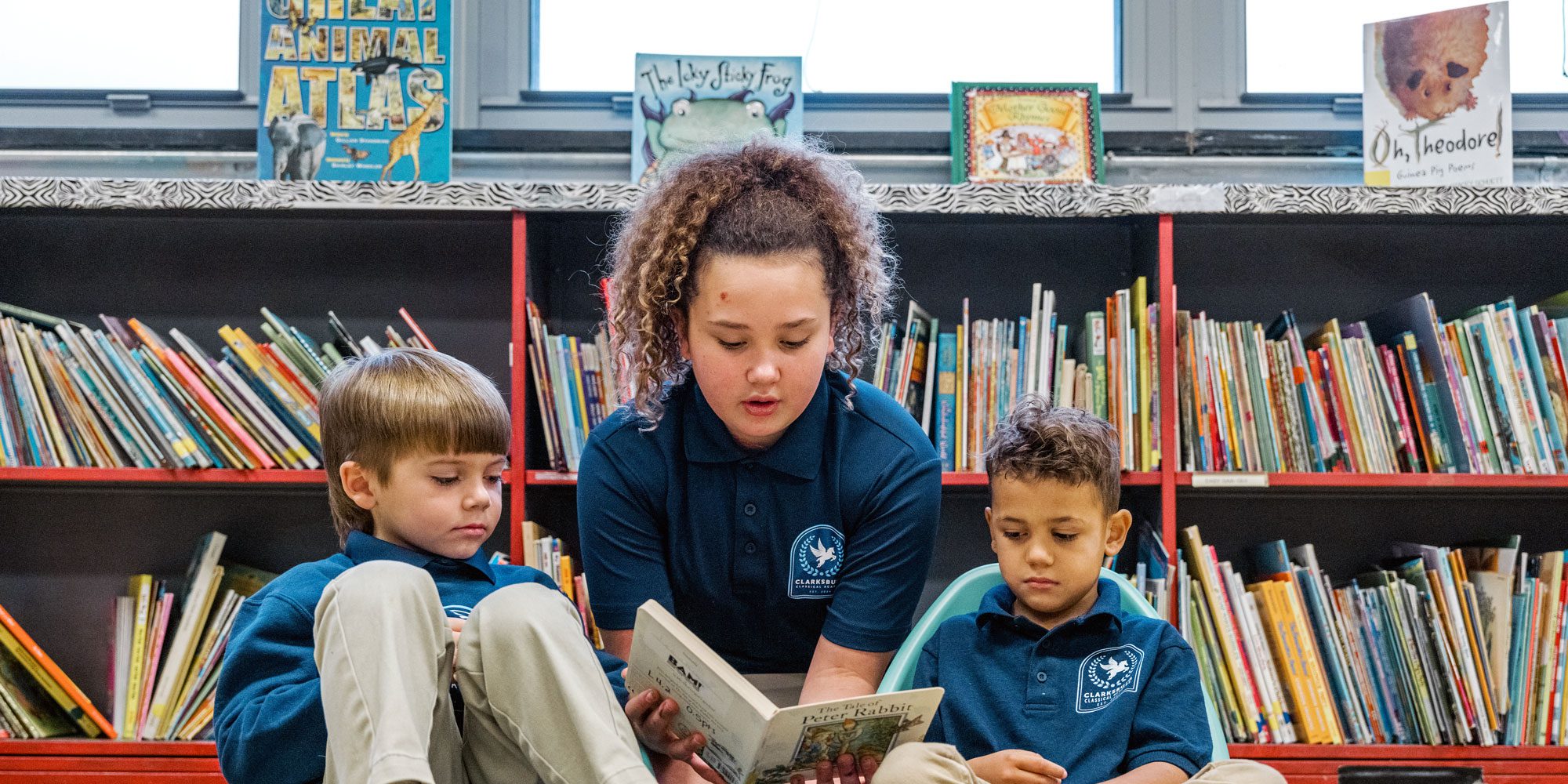 An older student reads a book aloud to two younger students in front of library shelves.