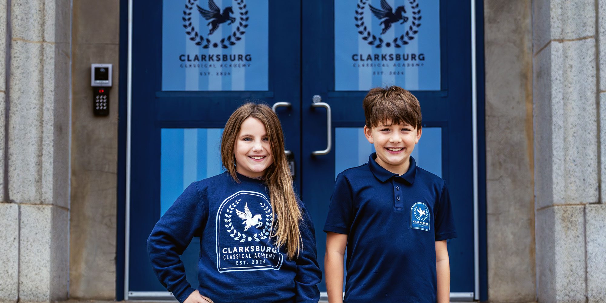 Two smiling Clarksburg Classical Academy students in front of the school's blue entrance doors.